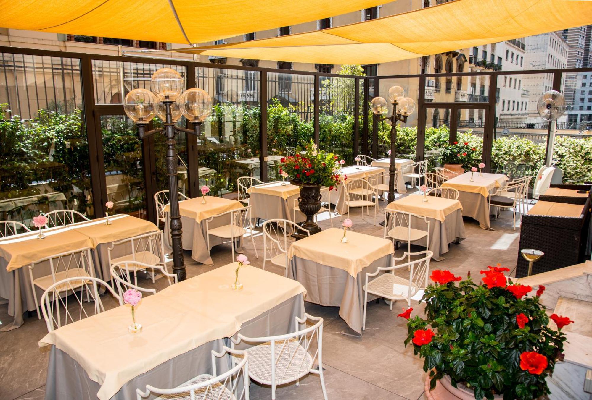 Hotel Bristol Palace Génova Exterior foto The photo shows an outdoor dining area, featuring several tables covered with light-colored tablecloths. Each table has a flower vase placed on it, adding a touch of decoration. The seating consists of white chairs, and the area is enclosed with glas