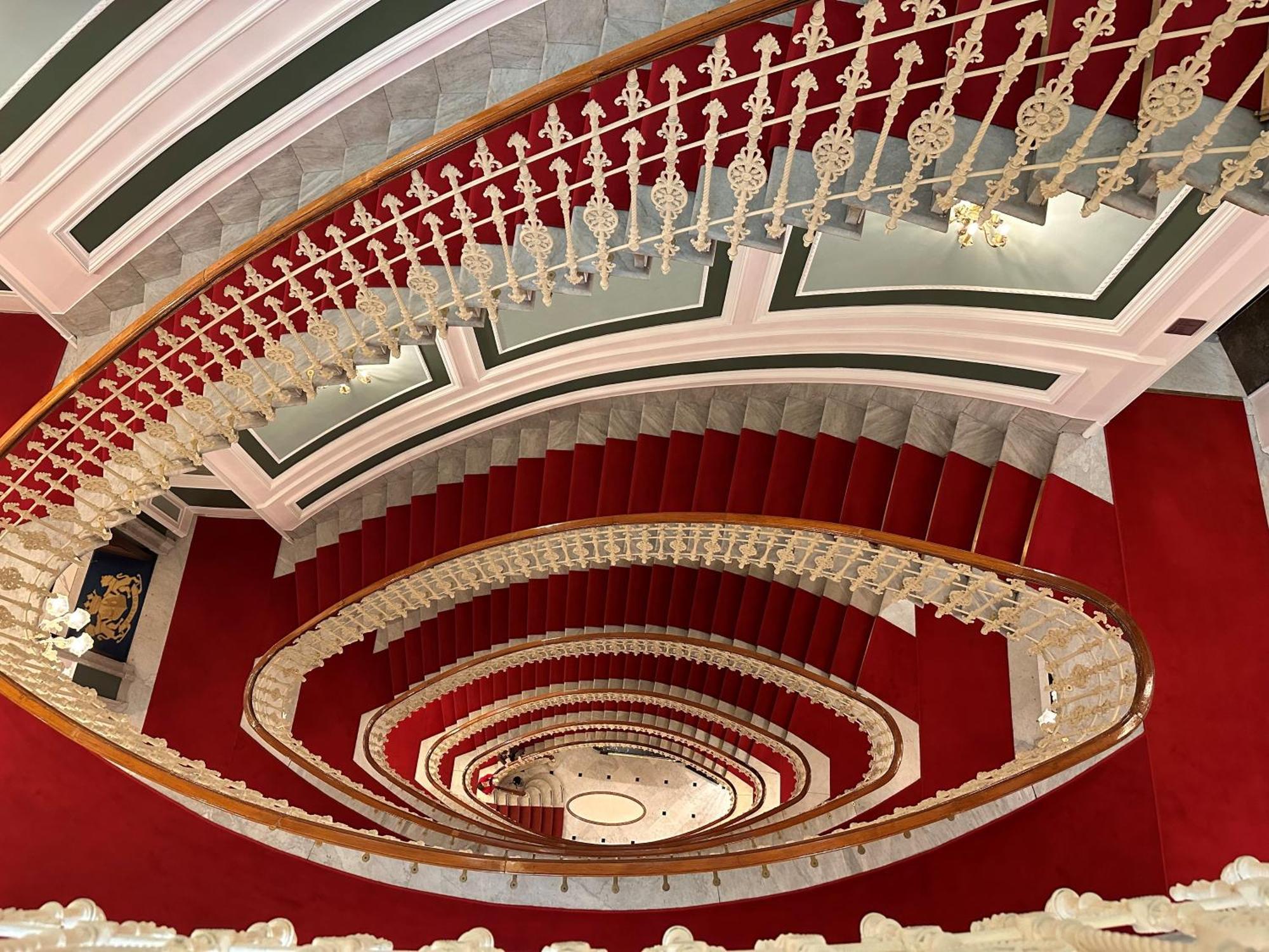 Hotel Bristol Palace Génova Exterior foto The photo shows a grand, spiral staircase viewed from above. The staircase features elegant white railings with intricate designs and is surrounded by a rich red carpet. The walls have a classic architectural style, with decorative elements in soft c