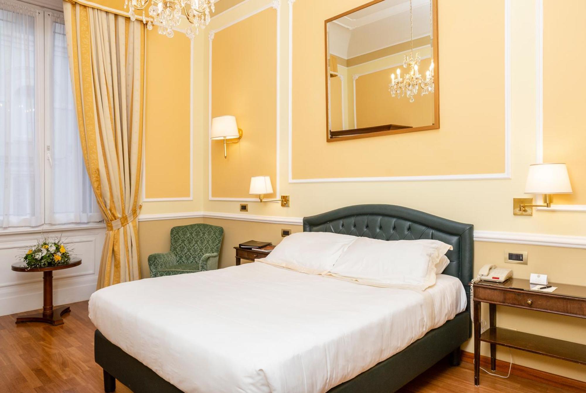 Hotel Bristol Palace Génova Habitación foto The photo shows a cozy hotel room with a warm color scheme. It features a large bed with a white duvet, neatly arranged. To the left, there's a light green armchair and a small table. The walls are painted a soft yellow, and there are elegant curtain