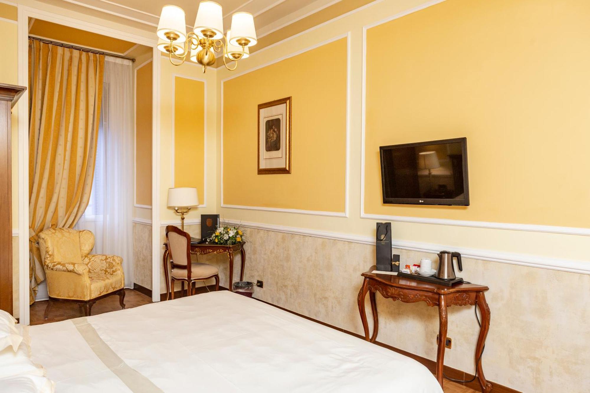 Hotel Bristol Palace Génova Habitación foto The photo shows an interior of a hotel room. The walls are painted a warm yellow color, and there's a chandelier hanging from the ceiling. On one side, there's a large window draped with elegant curtains. A comfortable armchair is situated in the cor