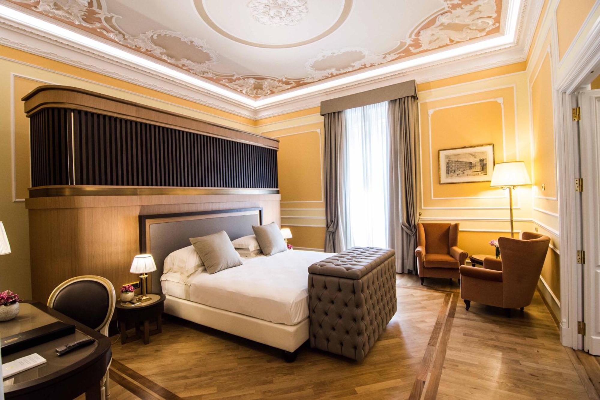 Hotel Bristol Palace Génova Exterior foto The photo shows an elegantly designed hotel room. It features a large bed with plush pillows, and a tufted ottoman at the foot of the bed. The walls are painted in a warm yellow tone, adorned with decorative molding. A pair of tall windows are draped